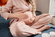 Pregnant woman in silk pajamas applying moisturizing cream on her belly before sleep or after shower while sitting on double bed
