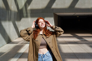 Canvas Print - Happy teen redhead girl hipster model standing near urban walls background wearing wireless headphones listening to music with eyes closed feeling joyful and relaxed outdoor.