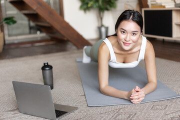 Wall Mural - Sporty asian girl in activewear doing workout at home, follow video sport guide, online fitness instructor on laptop, standing in plank on floor mat