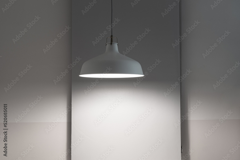 Pendant lamp hanging from the ceiling and giving spot light. Lamp next to the wall with window roller shades