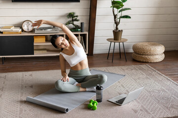 Wall Mural - Young asian fitness girl doing stretching, sport workout at home, stretch arms and looking at laptop, follow exercise video on computer