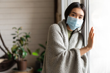 Wall Mural - Sick asian girl in medical face mask standing by the window and yearning to go outside, being on quarantine, ill with covid-19 or flu