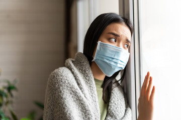 Wall Mural - Sick asian girl in medical face mask standing by the window and yearning to go outside, being on quarantine, ill with covid-19 or flu