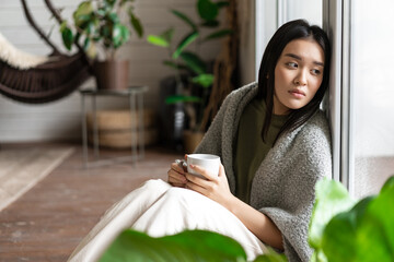 Wall Mural - Sad asian woman looking outside window, sitting on floor with miserable upset face, drinking coffee at home