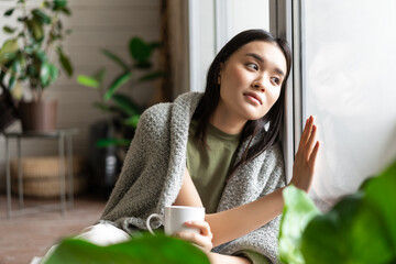 Wall Mural - Young woman on self quarantine at home, sitting by the window and touching glass, looking outside, drinking coffee