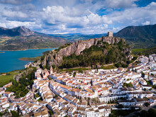 Aerial View Of Fortified Castle On Top Of Crag And White Residential Houses At Bottom Of Hill In Zahara De La Sierra On Background Of Turquoise Lake, Spain