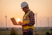 Portrait Of Engineer Wearing Yellow Vest And White Helmet Using A Computer Laptop On Site At Wind Turbines Field Or Farm, Sustainable Energy Concept