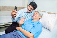 Indian Old Senior Patient In Hospital Bed Using Smart Phone Doing Video Call To Descendant Relatives With Son Sitting By Side - Elderly Medical And Healthcare Concept