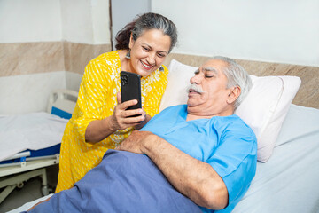 Wall Mural - Indian old senior patient in hospital bed using smart phone doing video call to descendant relatives with wife standing by side - Elderly medical and healthcare concept