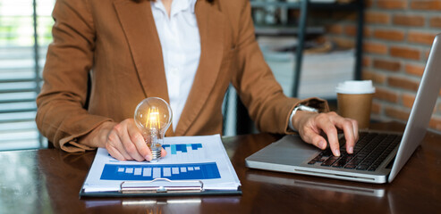 Wall Mural - Businesswoman holding and putting lightbulb on coins stack on table for saving energy and money concept..