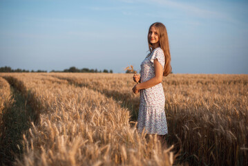 Poster - Side photo of a beautiful girl with blond hair holding spikelets of ripe rye looking to the camera. Portrait of a young girl walking in agricultural field on tractor traces