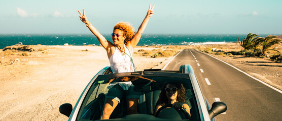 Travel with car vehicle and tourists summer destination concept. Happy and excited couple of women friends drive and have fun with a convertible and long straight road in background. Ocean and sea