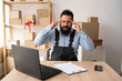 © Andrii Lysenko - Young Indian man working on laptop after packing the parcel for delivery. Small business owner working in office. Entrepreneur with headache thinking idea