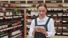 Young Manager Checks Shelves With Wine Bottles And Types On Tablet To Order Another Batch Of Alcohol. Woman Enjoys Working In Supermarket Closeup