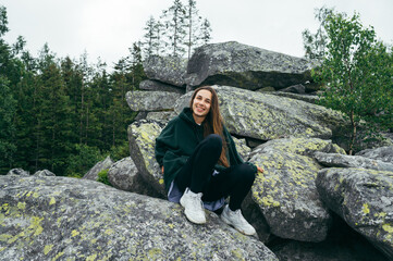 Attractive happy female tourist sitting on a rock in the mountains and posing for the camera with a smile on her face.