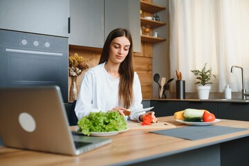 Wall Mural - Happy young woman making a salad at the kitchen, chopping vegetables, looking at laptop computer