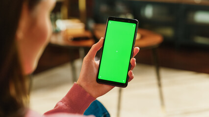 Fototapete - Feminine Hand Holding a Smartphone with Green Screen Mock Up Display. Female is Resting at Home, Watching Videos and Reading Social Media Posts on Mobile Device. Close Up Over the Shoulder Photo.