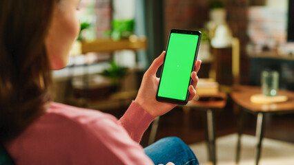 Fototapete - Feminine Hand Tapping on Display and Scrolling Feed on Smartphone with Green Screen Mock Up Display. Female Resting at Home, Checking Social Media on Mobile Device. Close Up Over the Shoulder Photo.