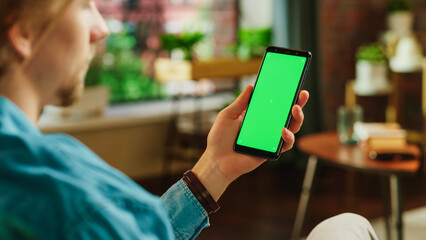 Fototapete - Man Holding a Smartphone with Green Screen Mock Up Display. Male is Resting at Home, Watching Videos and Reading Social Media Posts on Mobile Device. Close Up Over the Shoulder Photo.