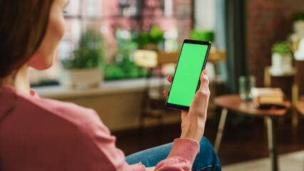 Leinwandbilder - Feminine Hand Holding a Smartphone with Green Screen Mock Up Display. Female is Relaxing at Home, Watching Videos and Reading Social Media Posts on Mobile Device. Close Up Over the Shoulder Photo.