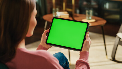 Fototapete - Female Holding Tablet Computer with Green Screen Mock Up Display. Woman Relaxing at Home, Watching Videos and Reading Social Media Posts on Mobile Device. Close Up Over the Shoulder Photo.