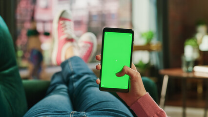 Fototapete - Feminine Hand Scrolling Feed on Smartphone with Green Screen Mock Up Display. Female is Relaxing on Sofa at Home, Watching Videos and Reading Social Media Posts on Mobile Device. Close Up POV Photo.
