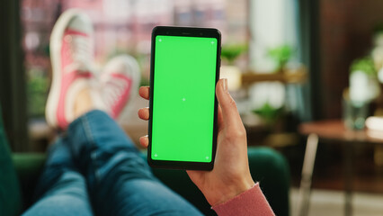 Fototapete - Feminine Hand Holding a Smartphone with Green Screen Mock Up Display. Female is Relaxing on a Couch at Home, Watching Videos and Reading Social Media Posts on Mobile Device. Close Up POV Photo.