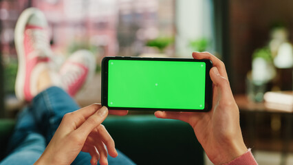 Fototapete - Woman Horizontally Holding a Smartphone with Green Screen Mock Up Display. Female is Relaxing on a Couch at Home, Watching Videos and Reading Social Media Posts on Mobile Device. Close Up POV Photo.