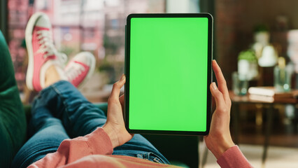 Fototapete - Feminine Hand Vertically Holding a Tablet with Green Screen Display. Female is Relaxing on a Couch at Home, Watching Videos and Reading Social Media Posts on Mobile Device. Close Up POV Photo.