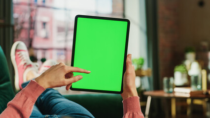 Fototapete - Feminine Hand Scrolling Feed on Tablet with Green Screen Mock Up Display. Female is Relaxing on Sofa at Home, Watching Videos and Reading Social Media Posts on Mobile Device. Close Up POV Photo.