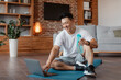 © Prostock-studio - Happy mature asian man in sportswear sitting on yoga mat with bottle of water, choosing fitness video, using laptop