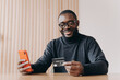 © VK Studio - Cheerful Aframerican man entrepreneur holding credit card and smartphone for online shopping