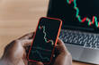 © VK Studio - Male hands of African man holds smartphone in front of laptop with trading charts on it
