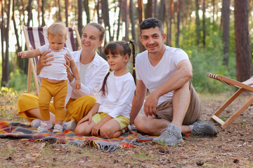 Wall Mural - Beautiful happy family enjoying moment while sitting on blanket in forest and playing with kids. Caucasian parents with small daughters relaxing in wood together.