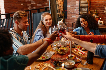 Diverse group of young people clinking glasses during dinner party in cozy warm setting