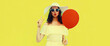 © rohappy - Portrait of beautiful young woman blowing her lips sending air kiss with red balloon wearing summer straw white hat on yellow background