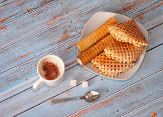 A plate with several different waffles, a cup of hot cappuccino and two sugar cubes on a wooden table.