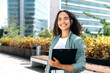 © Kateryna - Elegant confident young mixed race woman with curly hair, formally dressed, stands outdoors against the background of the business center, holding a folder with documents, looks at camera, smiles