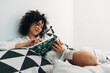 © Daniel - Happy young mixed race woman relaxes reading journal notes on bed. Copy space.