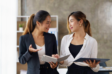 Wall Mural - Two beautiful Asian business women stand and talk about work using a tablet in the office.