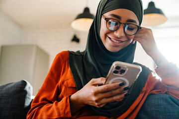 young beautiful smiling woman in hijab with phone on sofa