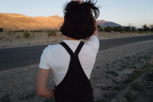 Back View Of Slim Person In Vintage Workwear Overalls With Short Hair Looking At Dusty Road In Rural Landscape