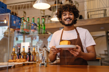 Young indian smiling handsome curly barista holding plate with cake