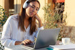 Young female student with headphones following an online course on laptop sitting at an outdoor cafe table