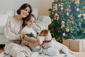 Happy winter holidays! Positive brunette woman embraces little girl pose with gifts on floor in room, jack russell terrier dog near, have fun near Christmas tree. Mom and daughter unpack boxes