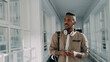 © silverkblack - Young handsome african american male student with big white headphones walking in long lighty glassy corridor of college holding smartphone looking into it texting someone and smiling.