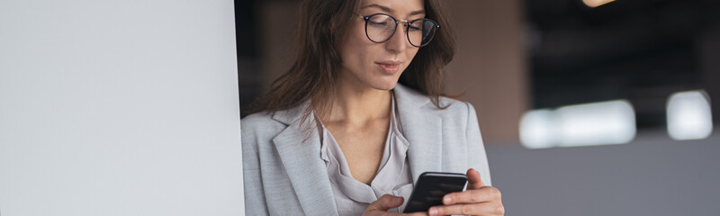 Businesswoman wearing glasses with mobile phone in hand standing in office. Blurred background