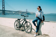 © PR Image Factory - stylish asian woman leaning on short white wall is having a coffee break from bike riding near golden gate bridge by the ocean on a sunny day in California USA.