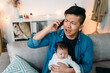annoyed asian dad holding a smpartphone is having an unpleasant conversation while taking care of his baby daughter on the couch in the living room at home.