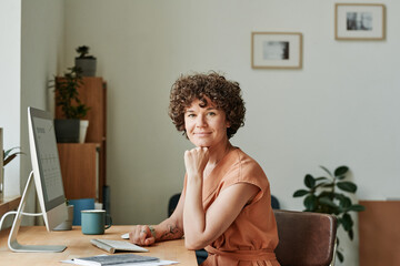 Portrait of young businesswoman sitting at her workplace in front of computer monitor at office and looking at camera